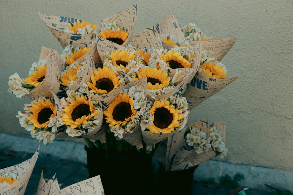 A bouquet of fresh sunflowers with vibrant yellow petals and dark brown centers, arranged with white daisies and greenery. The flowers are wrapped in decorative paper printed with the word 'LOVE' and 