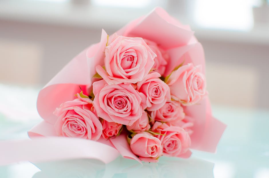 A bouquet of fresh pink roses arranged with tightly swirling petals and green stems, wrapped in soft pink tissue paper and tied with a pink ribbon, placed on a light-colored surface near a softly lit 