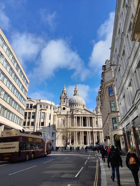 A view of St. Paul's Cathedral in London with its iconic dome and ornate façade, framed by surrounding historic and modern buildings on a clear day with blue sky and scattered clouds. The street in f