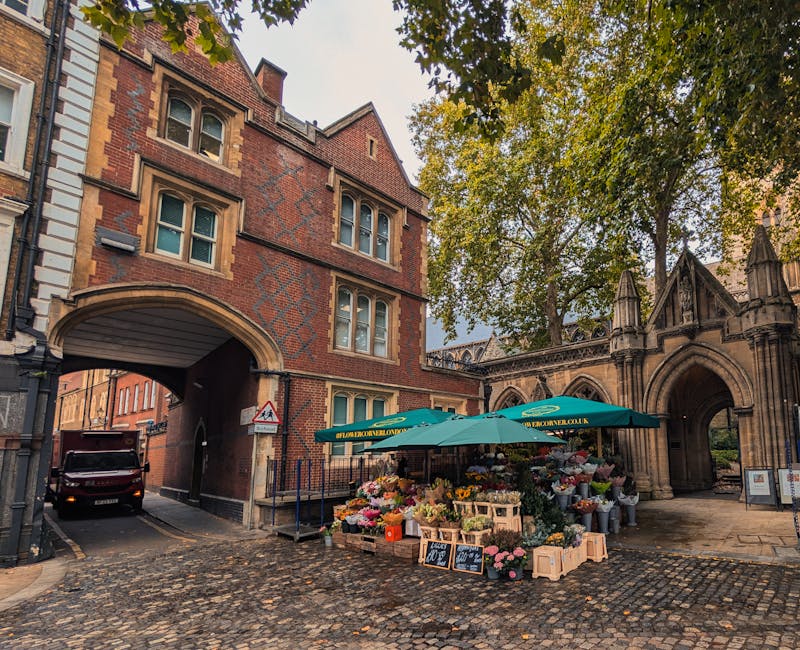 A flower stall situated on a cobbled street outside a historic brick and stone building with an arched gateway, featuring a green canopy and numerous wooden crates and boxes filled with a variety of f