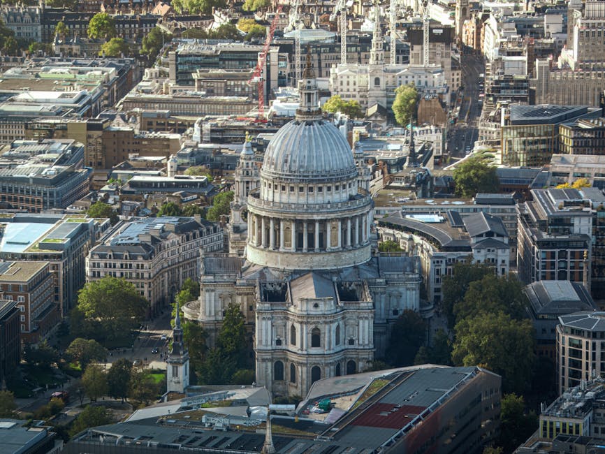 Aerial view of St. Paul's Cathedral in London, featuring its iconic large gray dome and classical architectural details. The cathedral is surrounded by modern and historic buildings, with trees lining