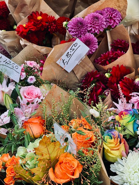 A colorful assortment of fresh flowers displayed in brown paper wrapping at a flower market or shop. The arrangement includes vibrant red, pink, purple, orange, yellow, and multicoloured roses, dahlia